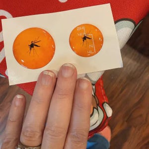 May include: Two orange circular stickers with a black mosquito illustration. One sticker has the mosquito illustration only, while the other has the illustration and additional white text and lines. The stickers are on a white background.