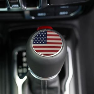 May include: A black gear shift knob with a red accent and a circular emblem featuring the American flag. The shift knob is set against a dark interior background, suggesting an automotive part.