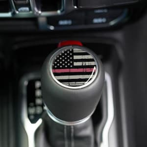 May include: A black gear shift knob with a distressed American flag design. The flag features white and black stripes, a field of stars, and a pink line. The knob is set against a dark interior.