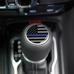 May include: Black gear shift knob with a graphic of the American flag with a blue stripe. The knob is round and has a red button on top. The background is the interior of a vehicle.