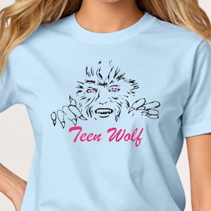 Pode incluir: Camiseta azul clara com uma ilustração em preto e branco de um lobisomem com olhos vermelhos e garras. O texto rosa em itálico diz "Teen Wolf". A camisa é feita de um material macio. Uma peça casual.