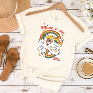 May include: A white t-shirt with a rainbow, a unicorn, and two cartoon characters riding on the unicorn. The text "Made in the 80's" is printed below the image.