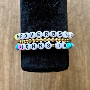 以下が含まれることがあります： 3本のビーズブレスレットの重ね付け。一番上のブレスレットは、PROVERBS 3:5と書かれた黒い文字入りの白い丸いビーズで構成されています。真ん中のブレスレットは小さなメタリックゴールドのビーズです。一番下のブレスレットは、JOHN 3:16と書かれた黒い文字入りの白いビーズと、その両端にカラフルな平らなディスク状のビーズが並んでいます。