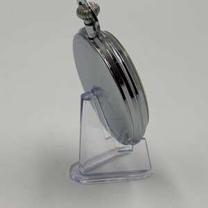May include: A silver pocket watch with a detailed, ridged crown and a circular face. The watch is displayed on a clear plastic stand, showcasing its polished metal finish. The watch's design is classic and elegant.