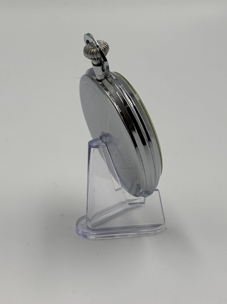 May include: A silver pocket watch with a detailed, ridged crown and a circular face. The watch is displayed on a clear plastic stand, showcasing its polished metal finish. The watch's design is classic and elegant.