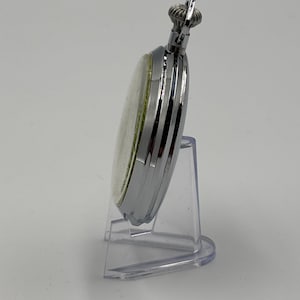 May include: A silver pocket watch with a white face and a textured crown. The watch is displayed on a clear plastic stand, showcasing its polished metal casing and classic design. A timeless accessory.