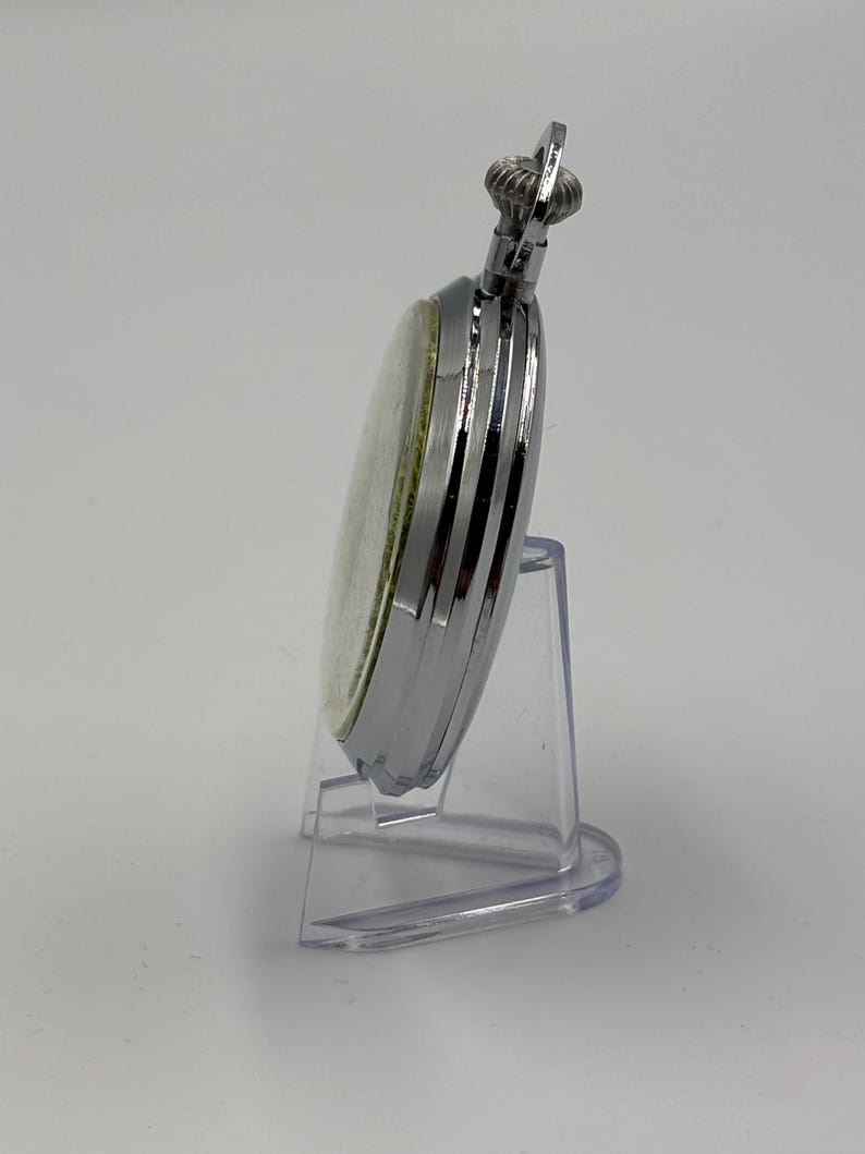 May include: A silver pocket watch with a white face and a textured crown. The watch is displayed on a clear plastic stand, showcasing its polished metal casing and classic design. A timeless accessory.