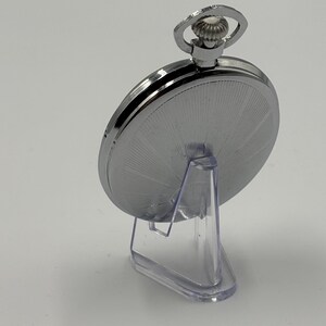 May include: A silver pocket watch with a detailed, radial design on the back. The watch is displayed on a clear plastic stand, showcasing its polished metal finish. The watch has a loop at the top for a chain.