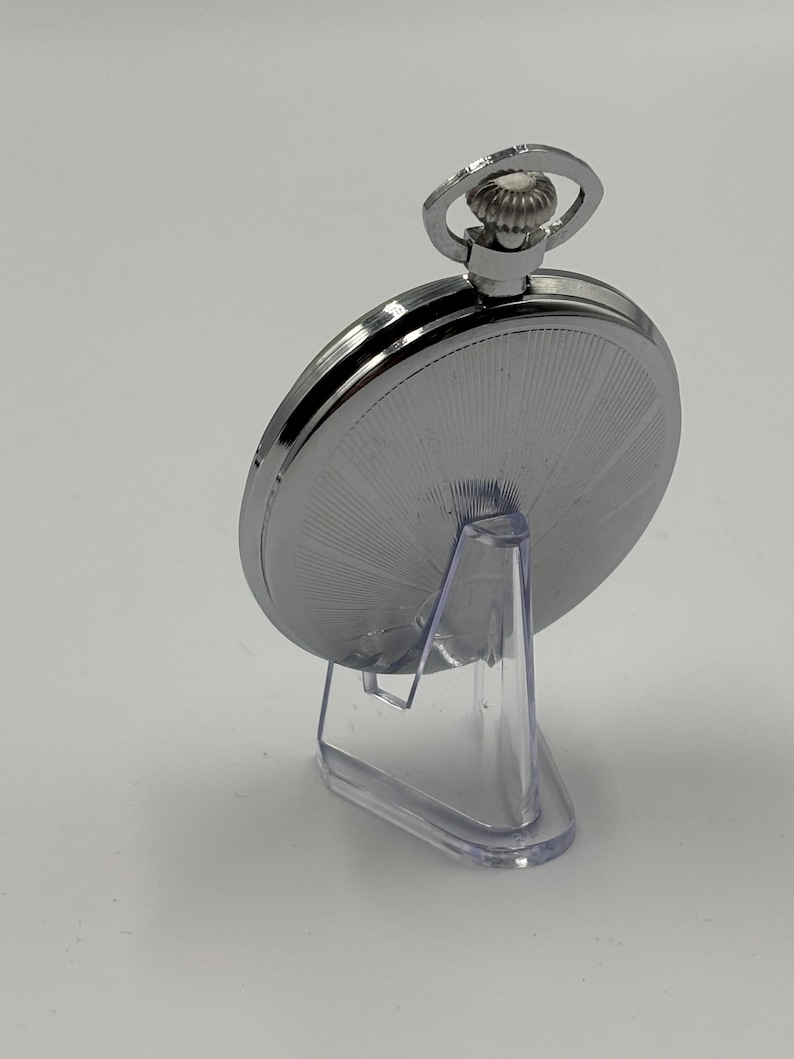 May include: A silver pocket watch with a detailed, radial design on the back. The watch is displayed on a clear plastic stand, showcasing its polished metal finish. The watch has a loop at the top for a chain.