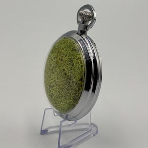 May include: A silver pocket watch with a clear face, revealing a textured green interior. The watch is presented on a clear plastic stand. The polished metal casing and the crown at the top are visible. The watch's design is intricate and detailed.