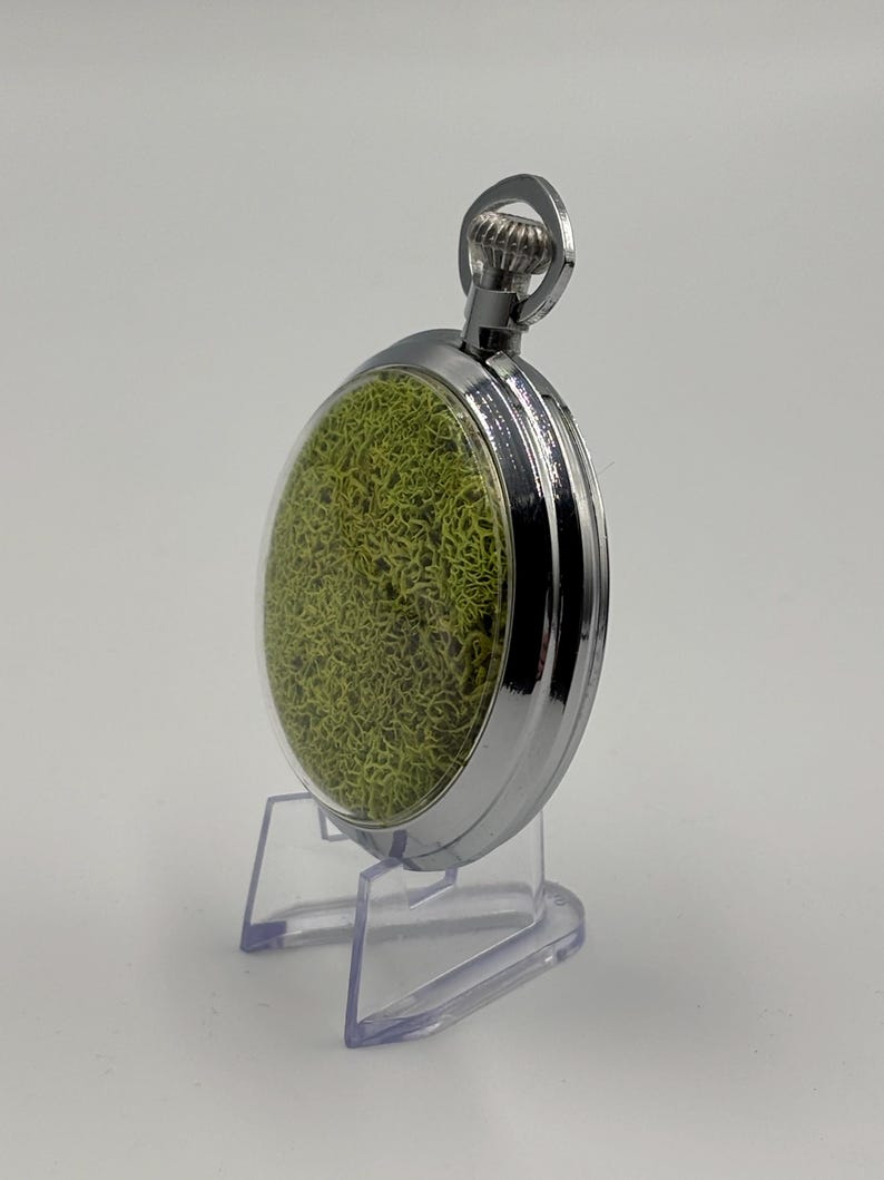 May include: A silver pocket watch with a clear face, revealing a textured green interior. The watch is presented on a clear plastic stand. The polished metal casing and the crown at the top are visible. The watch's design is intricate and detailed.