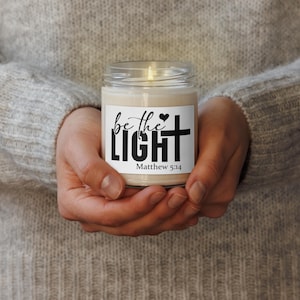 Puede incluir: Una vela encendida en un tarro de cristal transparente. La etiqueta dice "be the LIGHT" con una cruz y un corazón, y "Matthew 5:14". La vela se sostiene en manos ahuecadas, con un fondo neutro.