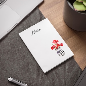 May include: A white notepad with the word "Notes" in cursive script. A watercolor illustration of red flowers in a striped vase is on the bottom right. The notepad is on a dark gray surface, near a laptop and a pen.