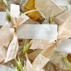 May include: Close-up of wedding favors with "Thank you" tags. Each favor is adorned with a peach-colored ribbon and a sprig of greenery. The tags also include the text "Belle & Anders".