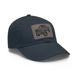May include: A navy blue baseball cap with a curved peak. A rectangular patch on the front displays a black graphic of a lifted truck. The cap is made of a soft fabric and has a classic, casual style.