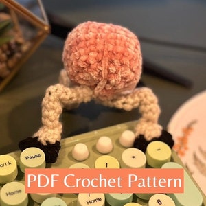 Op de afbeelding: Een roze gehäkelde hersenen met witte gehäkelde benen en armen, zittend op een groen toetsenbord met gele en witte toetsen. Het toetsenbord heeft de woorden "Home", "Pause" en "Scr Lk" op de toetsen. De afbeelding heeft de tekst "PDF Crochet Pattern" in een perzikkleurige doos.
