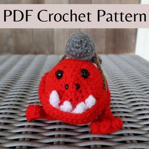 Op de afbeelding: Een handgemaakt rood gehaakt monster met witte tanden, zwarte ogen en een grijze bal bovenop. De tekst "PDF Crochet Pattern" wordt boven het monster weergegeven. Het monster zit op een wit vlechtwerk oppervlak.