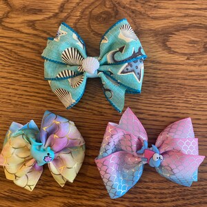 May include: Three handmade hair bows with a mermaid theme. The bows are made with blue, pink, and yellow fabric with a mermaid scale pattern. Each bow has a small 3D charm of a dolphin or fish.