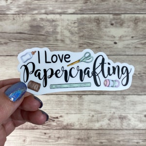 May include: White sticker with the text "I Love Papercrafting" in black. The sticker features illustrations of craft supplies, including scissors, a ruler, a spool of thread, and washi tape.