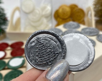 May include: A silver wax seal stamp with a design of a Christmas tree. The stamp is being held by a hand with a glittery nail polish.