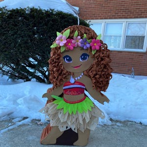 May include: A large, decorative cutout of a cartoon character with brown skin, curly brown hair, and a floral crown. The character wears a red and white top, a green skirt, and a necklace. The cutout is standing on a concrete surface with snow in the background.