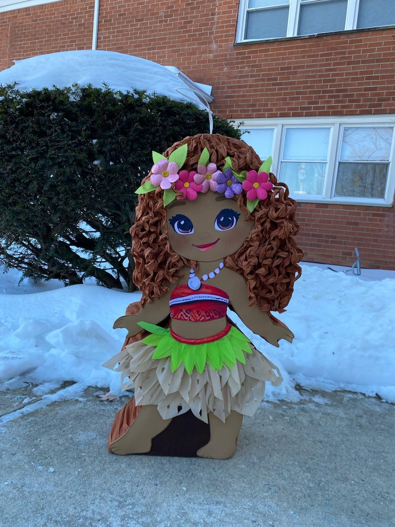 May include: A large, decorative cutout of a cartoon character with brown skin, curly brown hair, and a floral crown. The character wears a red and white top, a green skirt, and a necklace. The cutout is standing on a concrete surface with snow in the background.