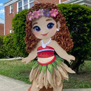 May include: A large, decorative cutout of a cartoon character with brown curly hair, a floral headband, and a white necklace. The character wears a red and white top, a green and tan skirt, and stands on a brown base. The background includes green bushes and a brick building.