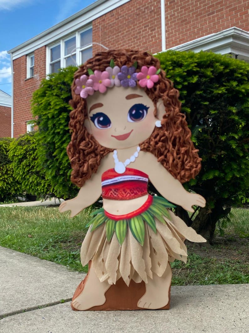 May include: A large, decorative cutout of a cartoon character with brown curly hair, a floral headband, and a white necklace. The character wears a red and white top, a green and tan skirt, and stands on a brown base. The background includes green bushes and a brick building.