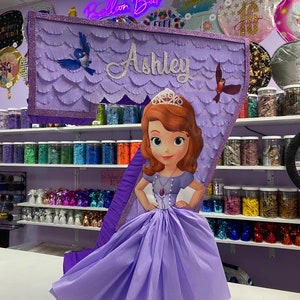 May include: Purple and silver number seven pinata with a princess character from the Disney movie Sofia the First. The pinata has the name "Ashley" written on it.