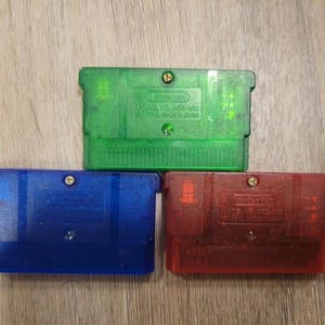 May include: Three Game Boy Advance cartridges in green, blue, and red. The green cartridge is at the top, with the blue and red cartridges below. The cartridges are made of translucent plastic and have a rectangular shape. The cartridges are on a wooden surface.