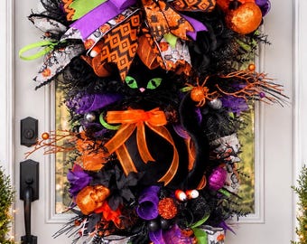 May include: A Halloween teardrop wreath featuring a central black cat with green eyes, surrounded by black mesh, orange and purple ribbons, patterned fabric with pumpkins and bats, orange glittered skulls, and black twig accents.