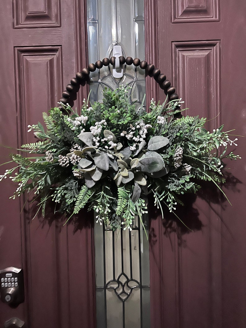 May include: A brown wooden beaded wreath with a green and white floral arrangement. The wreath is hanging on a dark red door with a glass panel.