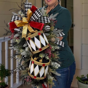 May include: A decorative holiday swag featuring two miniature drums with black and white diamond patterns, gold trim, and red accents. The arrangement includes black and white checkered ribbon, a gold glitter bow, pine branches, and red berries.