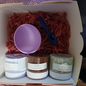 K&ouml;nnte beinhalten: Eine Geschenkbox gef&uuml;llt mit roten Papierschnipseln, einer lavendelfarbenen Sch&uuml;ssel und einem dunkelblauen L&ouml;ffel. Drei kleine Gl&auml;ser mit wei&szlig;en Etiketten sind sichtbar. Die Gl&auml;ser enthalten unterschiedlich farbige Inhalte, was auf ein Set von Bade- oder Sch&ouml;nheitsprodukten hindeutet.