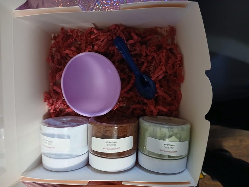 K&ouml;nnte beinhalten: Eine Geschenkbox gef&uuml;llt mit roten Papierschnipseln, einer lavendelfarbenen Sch&uuml;ssel und einem dunkelblauen L&ouml;ffel. Drei kleine Gl&auml;ser mit wei&szlig;en Etiketten sind sichtbar. Die Gl&auml;ser enthalten unterschiedlich farbige Inhalte, was auf ein Set von Bade- oder Sch&ouml;nheitsprodukten hindeutet.