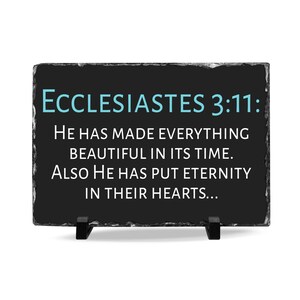 May include: A rectangular black slate plaque with the text "ECCLESIASTES 3:11: HE HAS MADE EVERYTHING BEAUTIFUL IN ITS TIME. ALSO HE HAS PUT ETERNITY IN THEIR HEARTS..." in white and teal lettering. The plaque is supported by two black stands.
