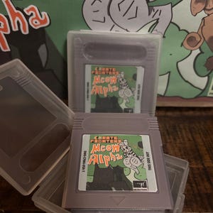 Könnte beinhalten: Zwei graue Nintendo Game Boy-Cartridges mit dem Spiel "Meow Alpha" werden in transparenten Kunststoffhüllen präsentiert. Das Spiel-Label zeigt eine Cartoon-Katze. Im Hintergrund befindet sich ein grünes Poster mit einer Katzenillustration.