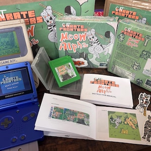 Könnte beinhalten: Eine Sammlung von Retro-Videospielartikeln, darunter ein blaues Game Boy Advance SP und ein klassisches Game Boy. Die Spiele tragen den Titel "Cat Karate Fighters: Meow Alpha". Die Verpackung und Handbücher zeigen Cartoon-Katzen-Illustrationen in Grün und Orange.