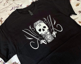 May include: Black t-shirt with a white graphic design featuring a skull, coffin, skeleton, and two crossed swords. The design includes the Roman numeral IX. The shirt is made of a soft material.