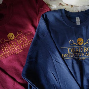 May include: Two long-sleeved shirts with the same logo. The logo is a skull with magnifying glasses and the text "Dead Boy Detective Agency est 1993". One shirt is maroon and the other is navy blue.