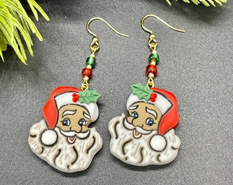 May include: A pair of festive Santa Claus earrings. Each earring features a Santa face with a red hat, white beard, and blue eyes. The earrings have gold-colored hooks and are adorned with red and green beads, and a green holly leaf.