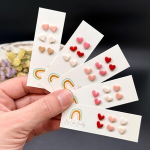 May include: A collection of heart-shaped stud earrings in various colors, including pink, red, and beige. The earrings are displayed on white cards with a rainbow design and the text "Joy Lake Jewelry".
