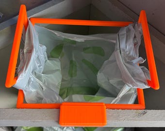 以下が含まれることがあります： 棚に取り付けられたオレンジ色のプラスチック製の袋ホルダー。ホルダーは、プラスチック製の買い物袋を保持するように設計されています。