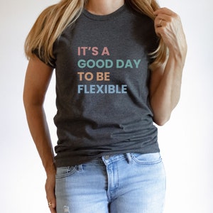 Puede incluir: Una camiseta gris oscuro con el texto "It's a good day to be flexible" impreso en colores pastel.