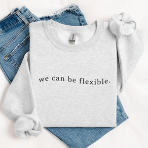 May include: A light gray sweatshirt with the phrase "we can be flexible." printed in black. The sweatshirt is folded on top of a pair of blue jeans. A white scrunchie is next to the sweatshirt.