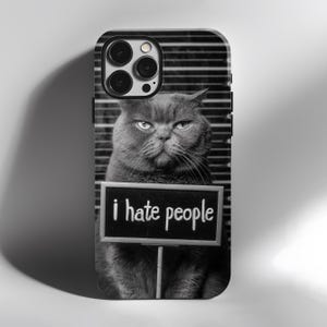 May include: Black and white phone case featuring a grumpy gray cat holding a sign that reads "i hate people." The background has a police lineup aesthetic. The case is designed for a smartphone.