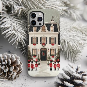 Op de afbeelding: Een telefoonhoesje met een besneeuwde winterscène met een wit huis, zwarte luiken en een krans op de deur. Rode strikken sieren de ramen en kerstbomen flankeren de ingang. Speelgoedsoldaten staan ervoor.