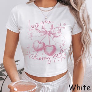 May include: White crop top with a pink cherry graphic and the text "Love you cherry much." The design features two cherries tied with a bow and star accents. The shirt is a casual, short-sleeved style.