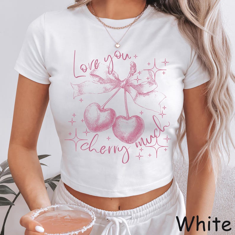 May include: White crop top with a pink cherry graphic and the text "Love you cherry much." The design features two cherries tied with a bow and star accents. The shirt is a casual, short-sleeved style.