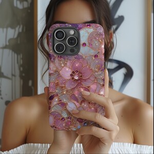 May include: A phone case with a floral design in shades of pink, purple, and gold. The case features a detailed pattern of flowers and gemstones, creating a sparkling, decorative look. The phone case is held in front of a person.